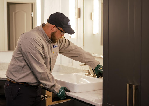 Professional plumber wearing safety glasses, gloves, and a branded Hamilton uniform adjusts the faucet of a modern bathroom sink. He is focused on his work, with water running from the faucet, set against a clean, well-lit bathroom with marble countertops and dark cabinetry.