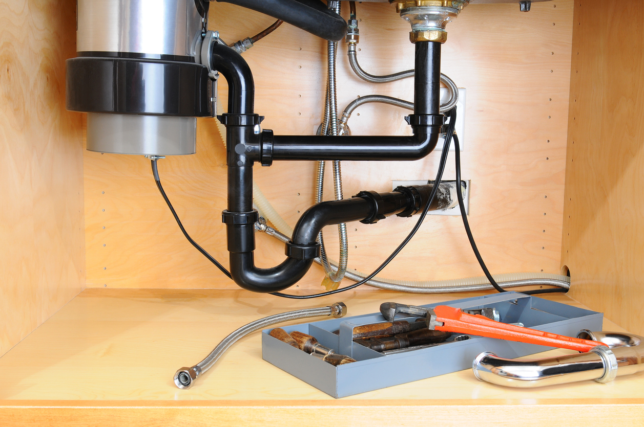 Garbage disposal and drain pipes beneath a kitchen sink with plumbing tools arranged for maintenance and clog prevention.